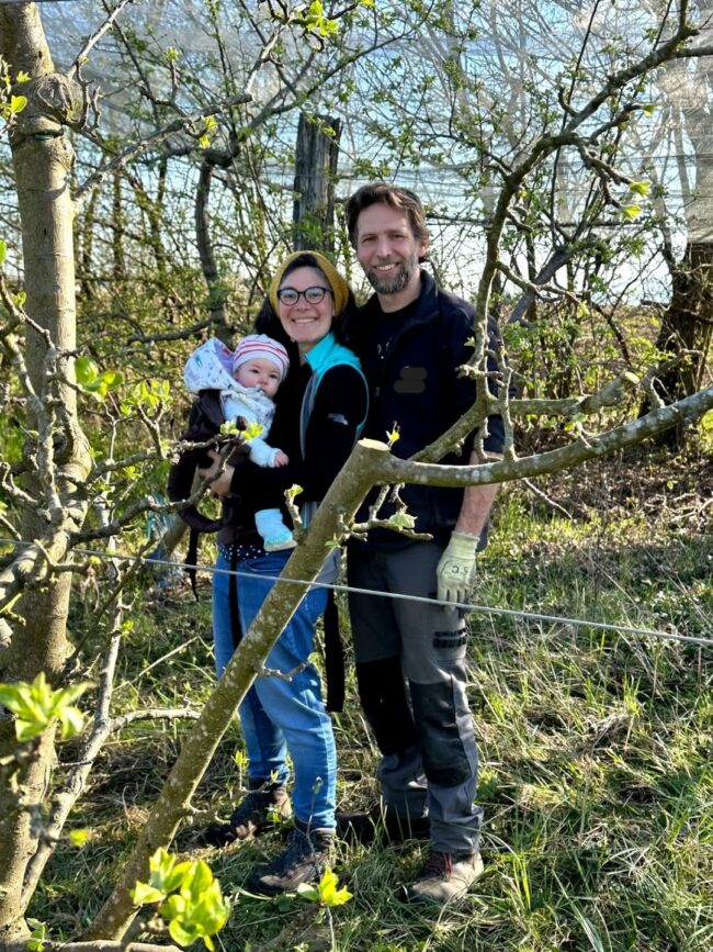 Mathilde et Daniel posent le sourire aux lèvres avec leur bébé lors d'un moment en famille dans le verger de leur ferme familiale
